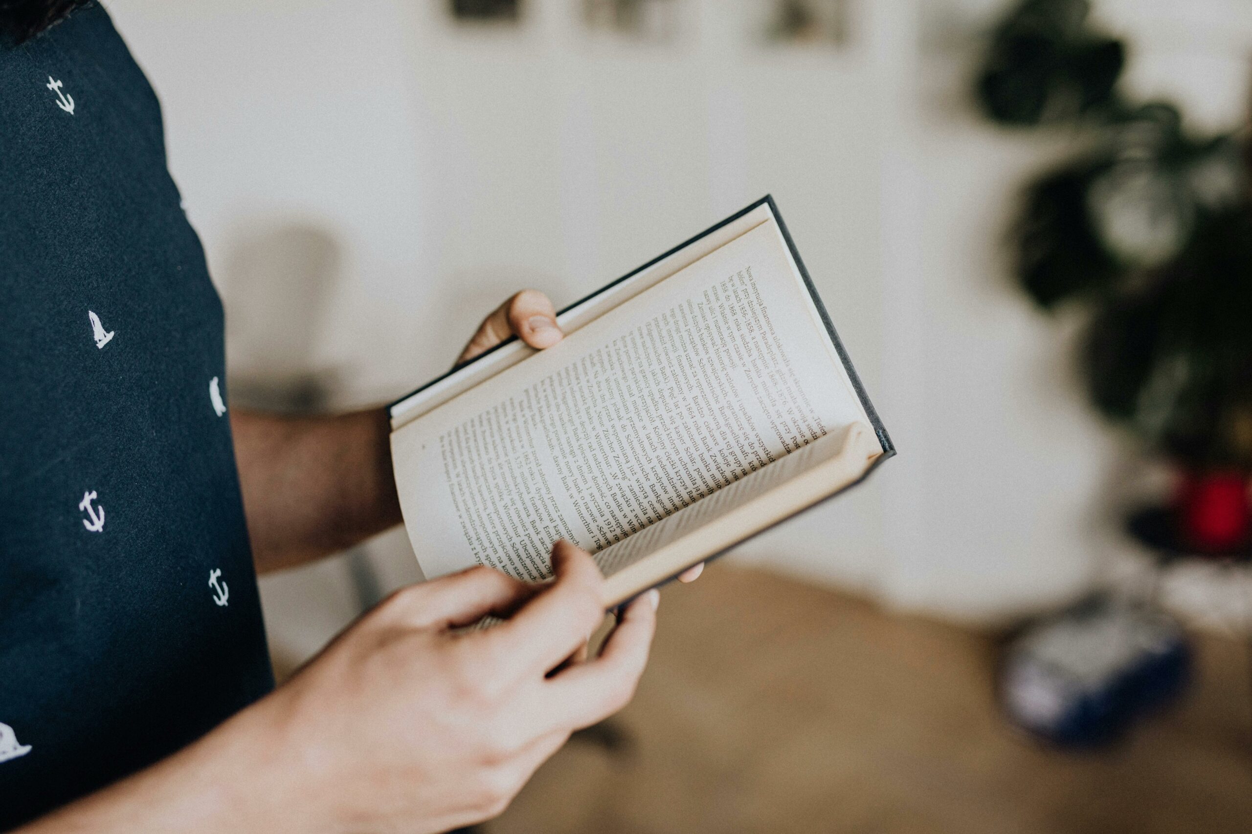 Close-up of a person holding and reading an open book indoors with a focus on hands and text.